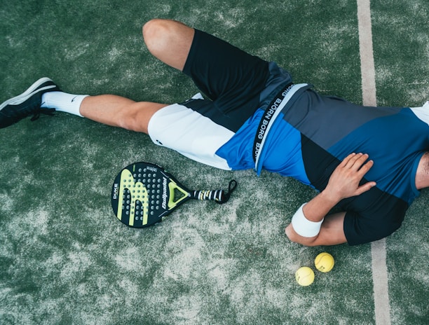 A person is lying on the ground, wearing athletic clothing that includes a blue and black top and white shorts. Nearby, there is a paddle racket and two yellow balls on a textured surface, which looks like a sports court. The person is wearing black sneakers and has a wristband on their arm.