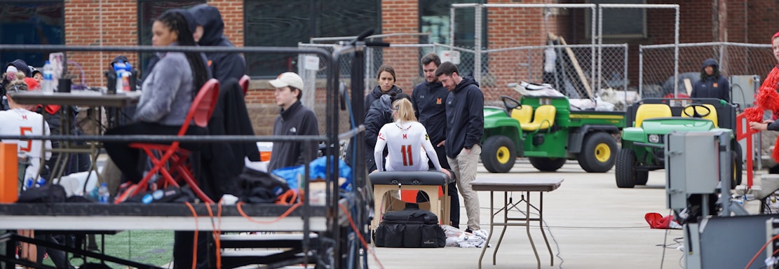 A group of people are gathered around an individual wearing a sports jersey with the number 11. They appear to be in a sports facility area, possibly a training or medical setup. There are tables, chairs, and equipment scattered around, with a brick building in the background. Some individuals are seated on a platform with electronics close by, suggesting a recording or monitoring activity. Utility vehicles and an orange barrier are present in the background.