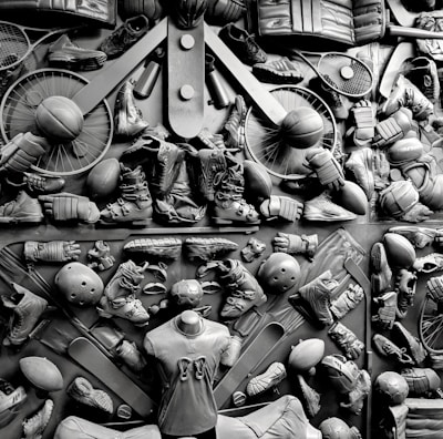 A complex arrangement of various sports equipment and attire, including tennis rackets, soccer balls, basketballs, helmets, shoes, gloves, and a mannequin torso wearing a T-shirt. The items are layered and artistically displayed in a monochrome color scheme, creating an intricate texture and visual pattern.