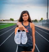 A young woman stands on a running track, holding a pair of white sneakers with orange details directly towards the viewer. She wears athletic clothing, including a sleeveless top with a letter 'L' on it and black shorts. The background features a sports field and a cloudy sky, suggesting a typical outdoor sports environment.