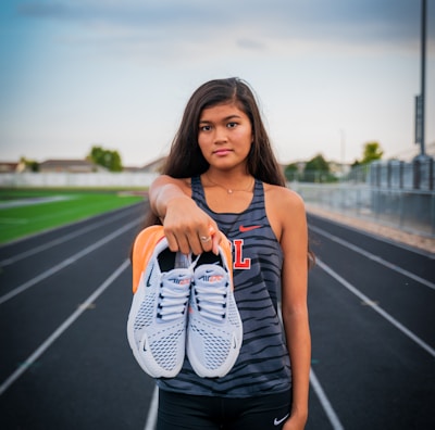 A young woman stands on a running track, holding a pair of white sneakers with orange details directly towards the viewer. She wears athletic clothing, including a sleeveless top with a letter 'L' on it and black shorts. The background features a sports field and a cloudy sky, suggesting a typical outdoor sports environment.
