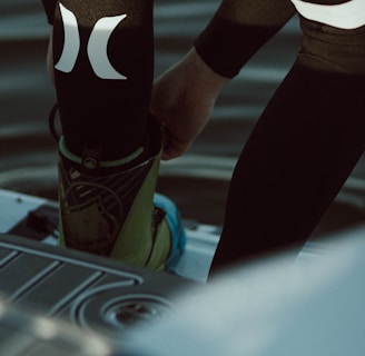 A person is putting on or adjusting water sports gear, standing on a platform above a body of water. The lower part of their body is visible, dressed in a black wetsuit with prominent white logos. Their footwear appears specialized, suggesting activities like wakeboarding or water skiing.
