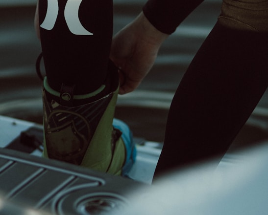 A person is putting on or adjusting water sports gear, standing on a platform above a body of water. The lower part of their body is visible, dressed in a black wetsuit with prominent white logos. Their footwear appears specialized, suggesting activities like wakeboarding or water skiing.