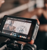 A digital camera screen displays a close-up of a person in athletic wear, possibly recording a fitness video. The person appears to be smiling and is in a well-lit space with a ring-like setting visible in the background.