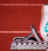 A close-up of a white athletic shoe with colorful speckles and a blue swoosh logo, resting on a starting block on a red track field. The shoe is positioned as if ready for a race, with a visible foot wearing a white sock inside the shoe.
