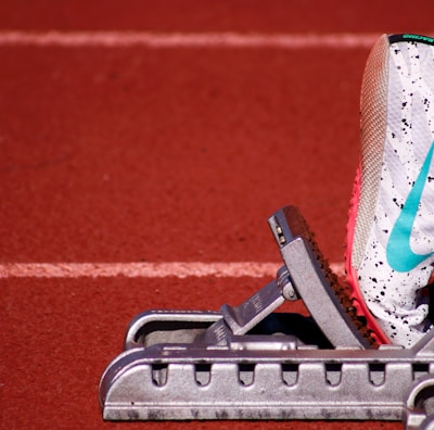 A close-up of a white athletic shoe with colorful speckles and a blue swoosh logo, resting on a starting block on a red track field. The shoe is positioned as if ready for a race, with a visible foot wearing a white sock inside the shoe.