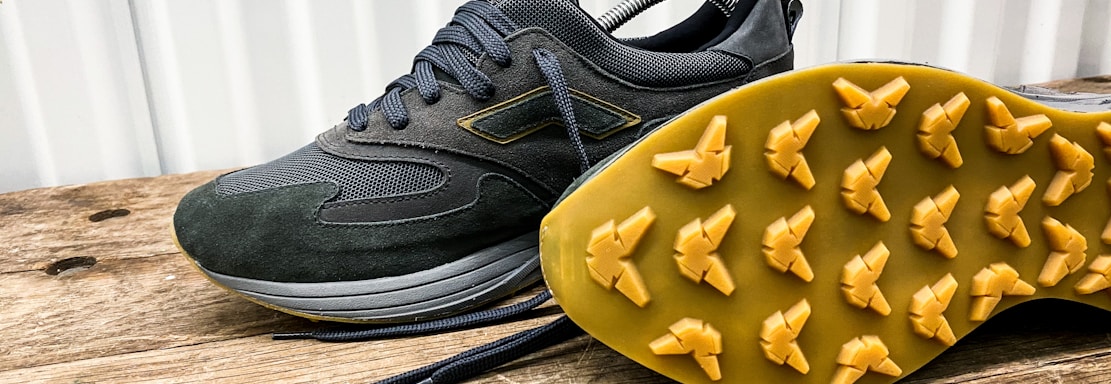A pair of dark-colored athletic shoes placed on a wooden surface, with one shoe laying on its side to display the sole. The shoes have a sleek design with a mix of suede and mesh materials, featuring a rubber sole with unique tread patterns for grip.