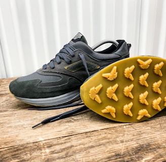 A pair of dark-colored athletic shoes placed on a wooden surface, with one shoe laying on its side to display the sole. The shoes have a sleek design with a mix of suede and mesh materials, featuring a rubber sole with unique tread patterns for grip.
