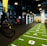 A modern gym space featuring a green track laid out on the floor with numbered lanes. TRX suspension trainers hang prominently in the foreground. The gym is equipped with various exercise equipment, including kettlebells and punching bags. In the background, there is a FIT sign near a reception desk under a low-ceilinged industrial design.