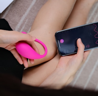 A person holds a bright pink wearable device in one hand while interacting with a smartphone in the other hand. The smartphone screen displays a graph with lines and numerical data, suggesting a tracking or monitoring application. The background consists of a soft, neutral-toned surface with a partial view of a box.