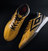 A pair of yellow and black sports shoes displayed against a dark background. The shoes feature a bold geometric pattern and are designed for athletic performance.