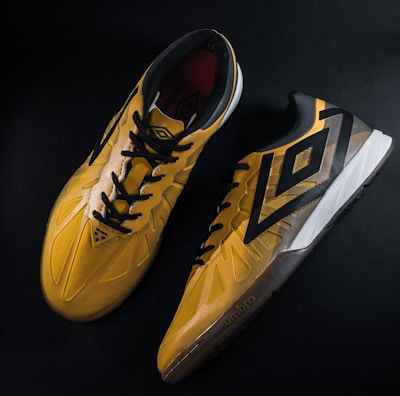 A pair of yellow and black sports shoes displayed against a dark background. The shoes feature a bold geometric pattern and are designed for athletic performance.