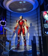 A high-tech suit displayed in a futuristic setting, illuminated by focused lighting. The surroundings include digital screens showing technical data and designs related to the suit. The environment appears to be a lab or exhibit with metallic and technological elements.