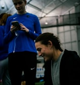 Two people are indoors in what appears to be a sports facility with a high ceiling and metal framework. One person is looking down at their phone with earphones in, wearing a blue long-sleeve athletic shirt. The other person is smiling and appears to be engaging with something or someone off-camera, dressed in darker athletic wear. The background is slightly blurred but shows additional people and possibly sports equipment.