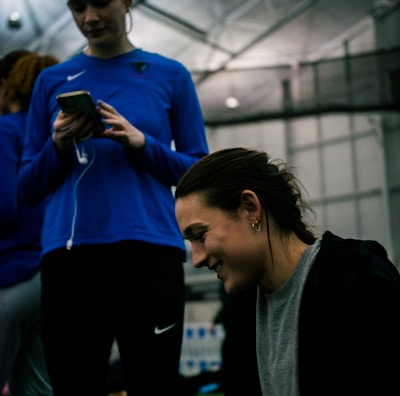 Two people are indoors in what appears to be a sports facility with a high ceiling and metal framework. One person is looking down at their phone with earphones in, wearing a blue long-sleeve athletic shirt. The other person is smiling and appears to be engaging with something or someone off-camera, dressed in darker athletic wear. The background is slightly blurred but shows additional people and possibly sports equipment.