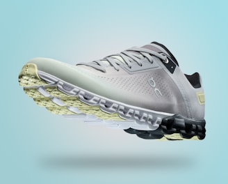 A single sports shoe is prominently displayed against a seamless, light turquoise background. The shoe features a sleek design with a gradient of light gray to dark gray tones, and is equipped with a distinctive CloudTec sole for cushioning. The branding logo is subtly visible on the side.