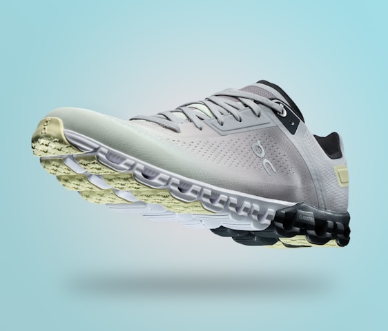 A single sports shoe is prominently displayed against a seamless, light turquoise background. The shoe features a sleek design with a gradient of light gray to dark gray tones, and is equipped with a distinctive CloudTec sole for cushioning. The branding logo is subtly visible on the side.